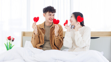 Asian young couple love emotions shine happy enjoy smiling show holding red heart shape paper sitting on white bed at home together relationship lifestyle surprise romantic celebrating Valentine day.