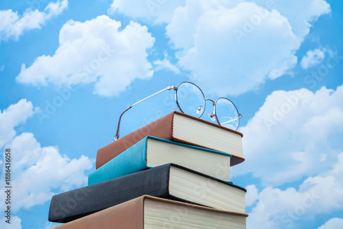 Stack of books with eyeglasses against a blue sky with clouds. Education, knowledge, learning, academic growth concept.