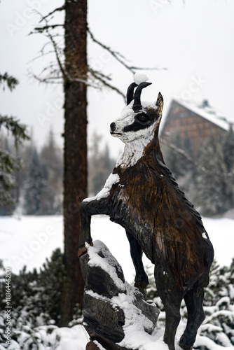  A detailed wooden sculpture of a mountain goat standing on a snow-covered rock. In the blurred background, a large alpine hotel and evergreen forest create a winter resort scene.