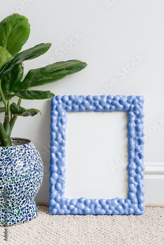 Blank vertical bubble-textured frame painted ultramarine blue, standing on floor in modern living room with neutral sofa, minimalist conceptual interior, soft diffused daylight