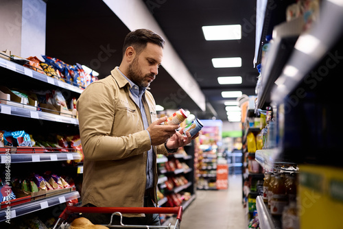 Fotografía Mid adult man hesitating while choosing baby food in supermarket.