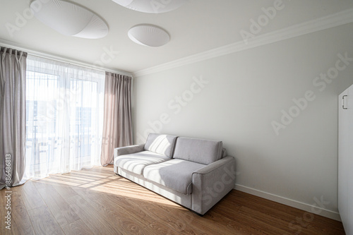 minimalist living room with a light grey sofa, wooden flooring, sheer white curtains, large window, and modern ceiling lights; clean white walls, bright and airy.