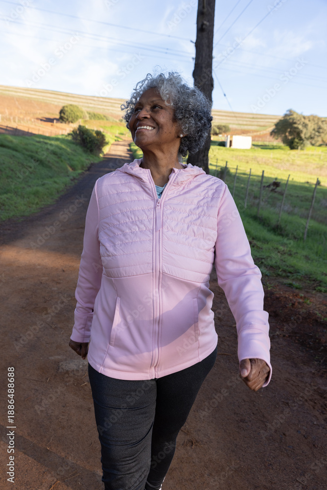 Naklejka premium African american senior woman walking dirt road wearing pink jacket past vineyard rows