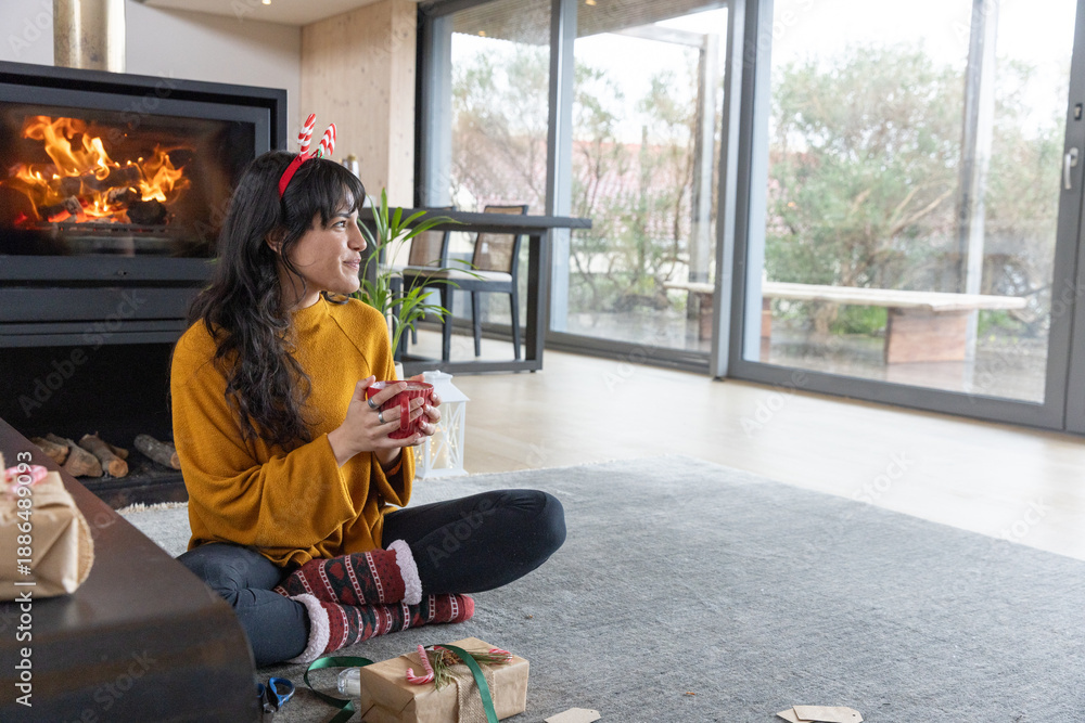 Fototapeta premium Woman sitting cross-legged on rug in living room holding mug among wrapped gifts and fireplace