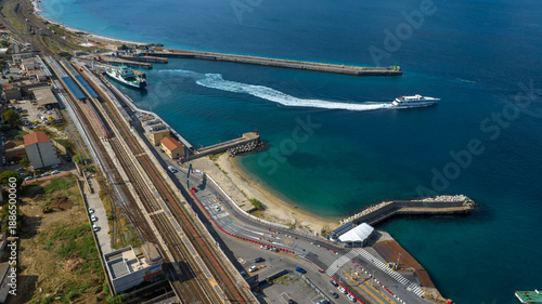 High-angle aerial view of a white hydrofoil leaving a white wake in a harbor. The scene includes railway tracks and a small sandy beach. It is the port of Villa San Giovanni, in Calabria, Italy.