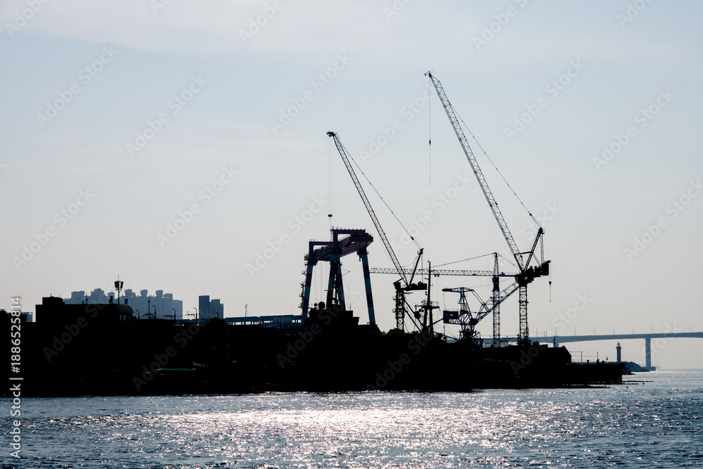 Obraz premium Silhouette heavy industrial cargo vessel and high-rise building, trawlers docked in the harbour while loading containers via cranes under a sunny sky in commercial Busan port city, South Korea