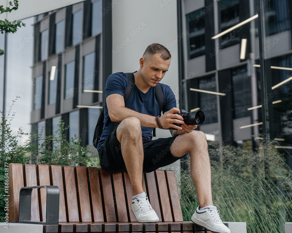 Fototapeta premium Young photographer sitting on a modern wooden bench outside a glass office building, holding a mirrorless camera; casual summer outfit, urban lifestyle portrait.