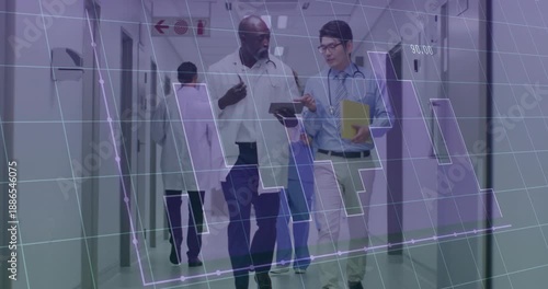 Doctor raising tablet toward physician, swiping graphs for review while nurse moving down corridor