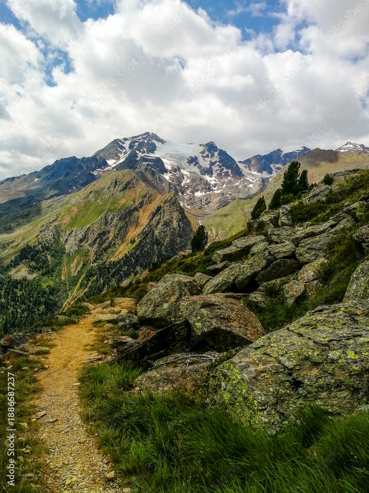 Obraz premium Vertical View of a Narrow Alpine Trail Along a Rocky Ridge with Snowy Peaks, Dramatic Clouds and Tree Line in the High Mountains of Trentino Alto Adige, Italy