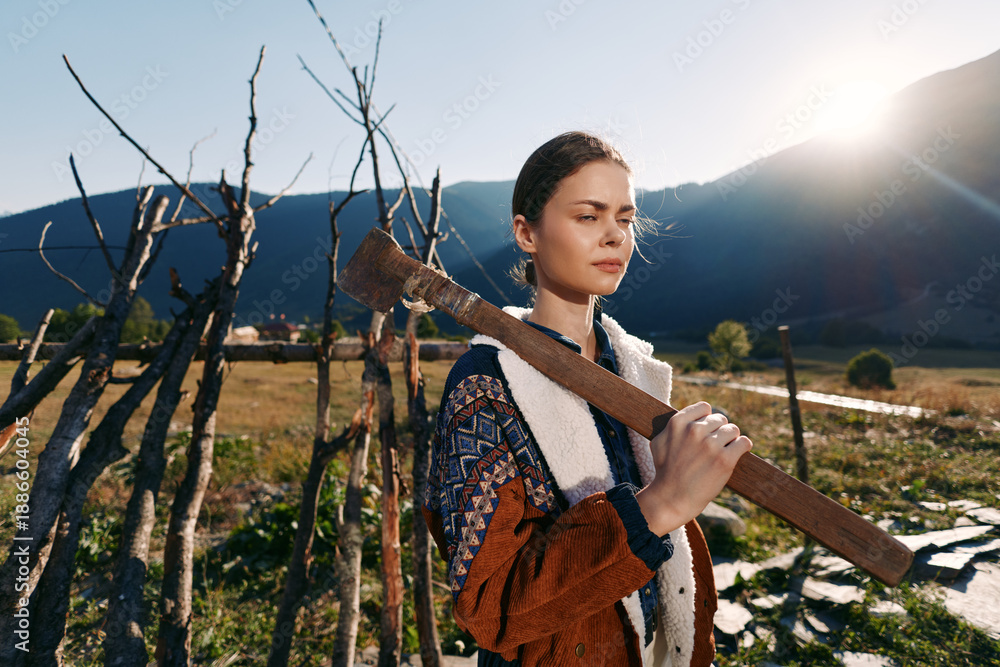 Fototapeta premium Woman with axe on her shoulder in a countryside farm setting by mountains at sunrise, portrait of a young rural worker in sweater carrying tool outdoors in golden sunlight.