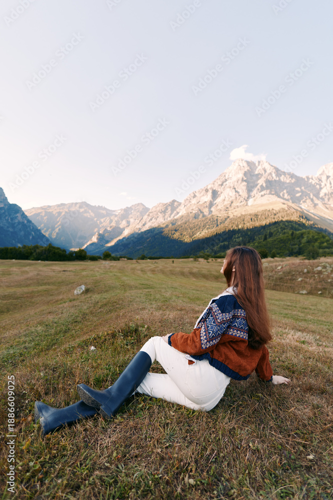 Fototapeta premium woman in sweater and boots sitting in meadow, mountains landscape and nature around her: young female with longhair relaxes on grassy field at dusk, enjoying serene outdoor solitude and scenic view