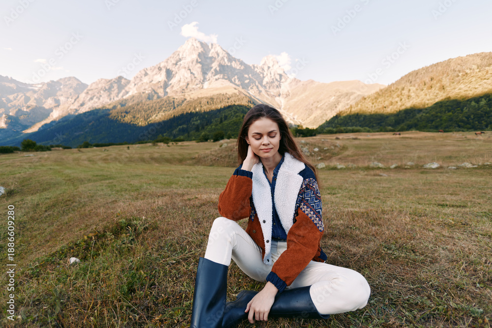 Fototapeta premium Woman portrait sitting in meadow with mountains in background, wearing shearling jacket and rubber boots, autumn outdoor scene, peaceful nature, relaxed young traveler.
