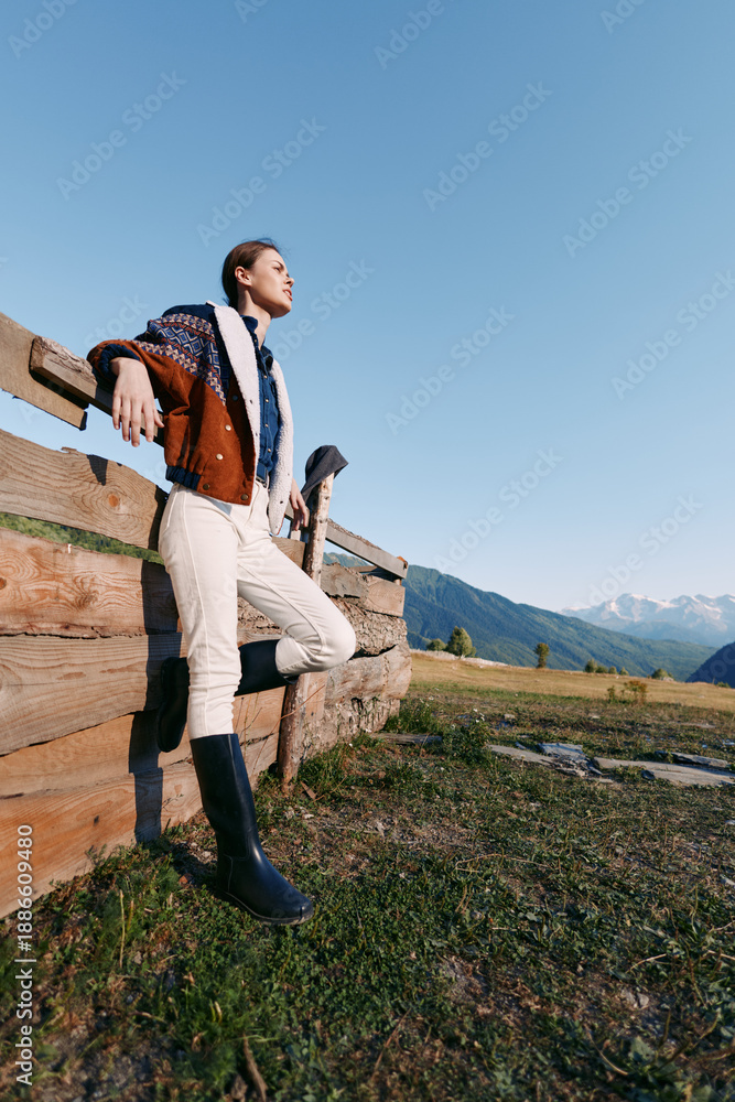 Fototapeta premium Woman leaning on wooden fence in countryside mountains, portrait outdoors wearing jacket and tall boots, looking into the distance during travel and nature lifestyle.