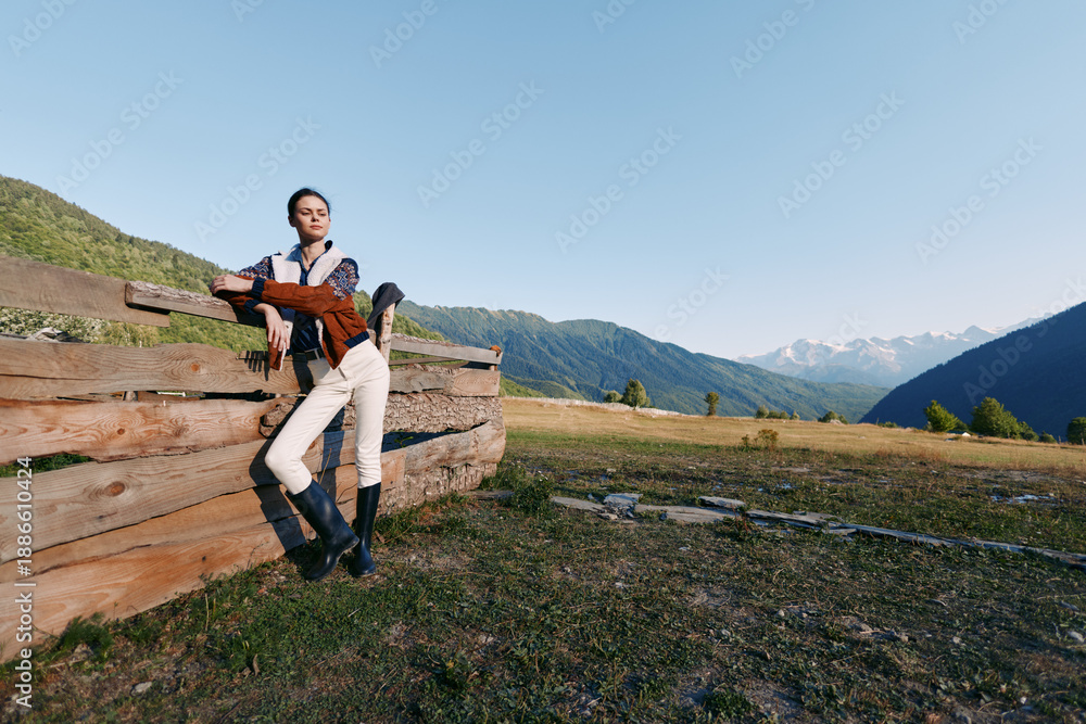 Fototapeta premium Woman leaning on wooden fence in countryside meadow with distant mountains, wearing riding boots and casual jacket, portrait lifestyle outdoors in wide open field under clear sky.