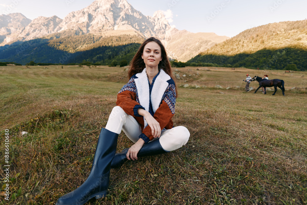 Fototapeta premium Woman sitting in mountain meadow wearing rubber boots and sweater, portrait with distant horse and rider, wide landscape view, outdoor nature relaxation and scenery.
