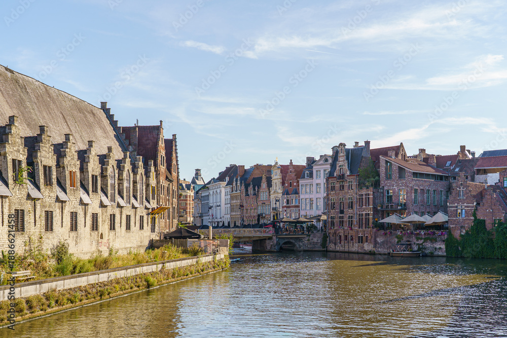 Fototapeta premium Buildings of varying shapes and colors line the riverbank in Ghent, Belgium. The large brick building is on the left. A bridge over the river is visible
