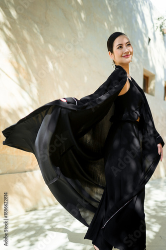 A full-length smiling woman dressed in a black abaya is captured in natural light. Traditional Arab-Muslim clothing.