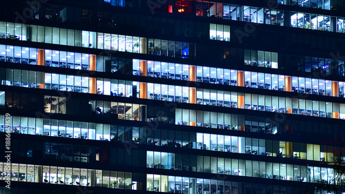 Big glowing windows in modern office building at night, in rows of windows light shines. Fragment of the glass facade of a modern corporate building at night. Modern glass office  in city.