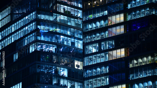 Big glowing windows in modern office building at night, in rows of windows light shines. Fragment of the glass facade of a modern corporate building at night. Modern glass office  in city.