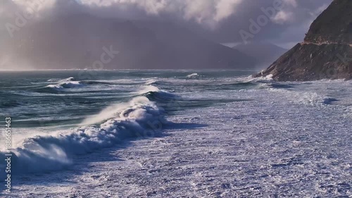 Aerial view of the coastline with waves crashing onto the shore under a cloudy sky, creating a dynamic interplay of light and shadow, Cape Town, Western Cape, South Africa.