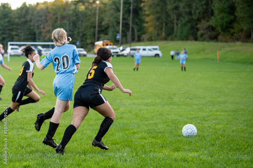 Obraz premium Female high school athletes from rival teams chase a soccer ball on the field in a dynamic action image of youth sports, competition, teamwork, and athletic determination.