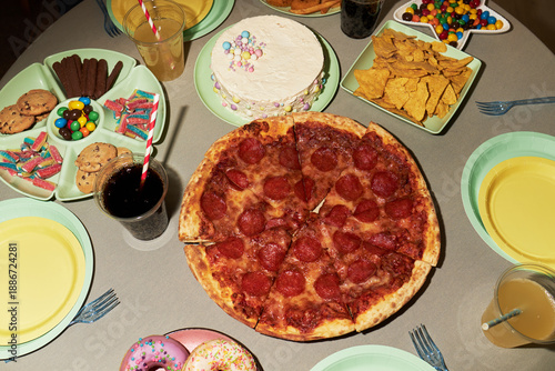 Overhead view showing pepperoni pizza, cake, cookies, donuts, tortilla chips, candy, and soft drinks arranged on table with empty plates and cups prepared for group gathering