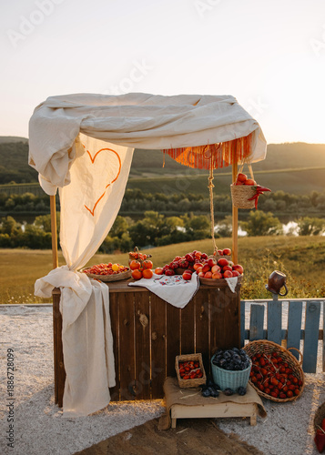 Rustic outdoor fruit market stand at sunset with apples, tomatoes, and grapes, styled for a countryside wedding or event. Romantic farm aesthetic with natural light.
