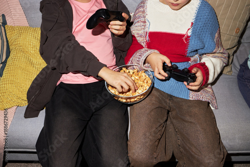 Two children sitting on sofa holding video game controllers and sharing bowl of popcorn, one child reaching for snack while both focusing on playing video game together