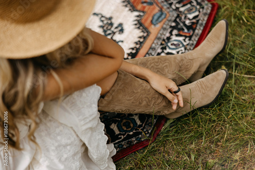 Woman wearing suede western boots sitting on a patterned rug outdoors, relaxed summer lifestyle scene with natural textures, grass, and boho-inspired fashion details.