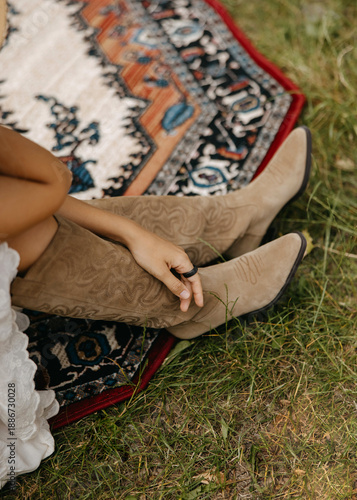 Boho dressed woman wearing beige suede cowboy boots while sitting on a patterned rug outdoors. Rustic detail with western style, natural light, and relaxed countryside vibes.