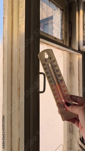 Thermometer in a woman's hand against the background of an old wooden window with frosty patterns. The concept of cold snaps, frosts. High humidity in the room.