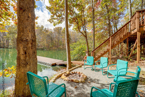 View of turquoise chairs encircle a stone-ringed fire pit near a tranquil lake reflecting the vibrant autumn foliage in Hamden, Ohio, United States.