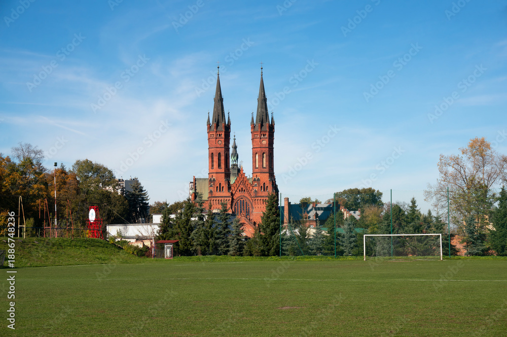 Obraz premium Playing field, football goal. Church of Holy Family (kościół Świętej Rodziny) in background, brick Neo-Gothic two-towered temple was built between 1904 and 1906. Tarnów, Poland.