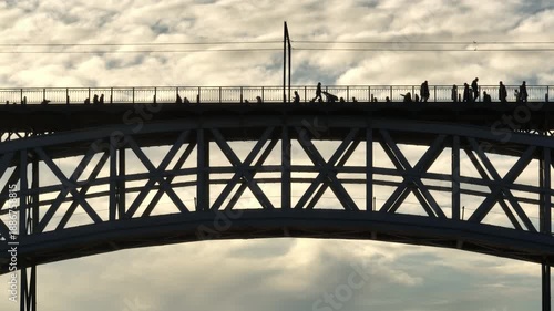 Silhouetted pedestrians people walking on the upper deck of Dom Luis I Bridge, spanning the Douro River in Porto, Portugal, at sunset, showcasing the iconic architecture and busy urban life