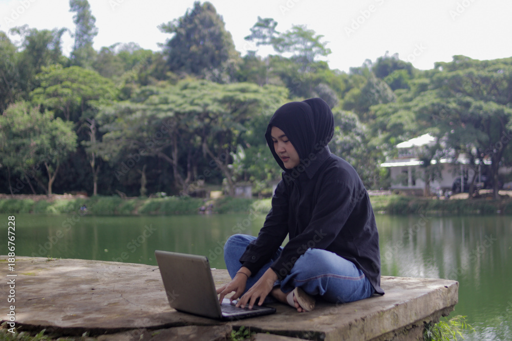 Fototapeta premium Young Asian woman wearing a hijab works on a laptop beside a lake in an outdoor setting. Remote work lifestyle, digital nomad, freelance work, online business, studying, and flexible workspace concept