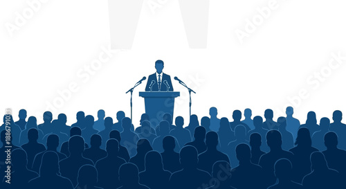 A speaker stands at a podium addressing a large, attentive audience in a dimly lit room with spotlights overhead.