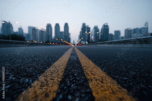 Low angle view of an asphalt road with yellow lines leading towards city buildings. It is suitable for illustrating urban planning, transportation, and infrastructure.