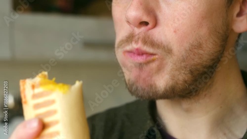 Extreme close up young caucasian bearded man with great appetite biting, chewing french hot dog, grilled baguette with sausage and mustard sauce, showing his satisfaction. Man eat french hot dog
