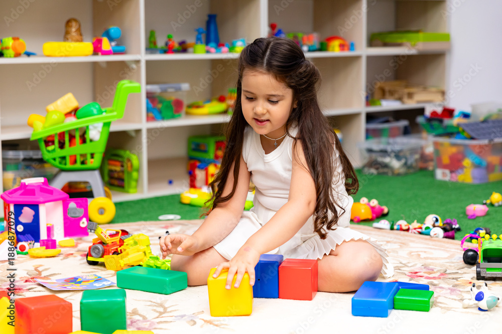 Fototapeta premium Little girl with long dark hair stacking colorful building blocks on a soft rug in a bright playroom filled with toys and storage shelves. Childhood and entertainment concept. High quality photo