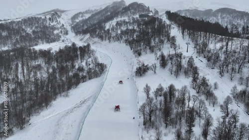 Cinematic drone shot of red snow grooming machines preparing the ski runs for tourists in the Bakuriani mountain resort during the winter season.