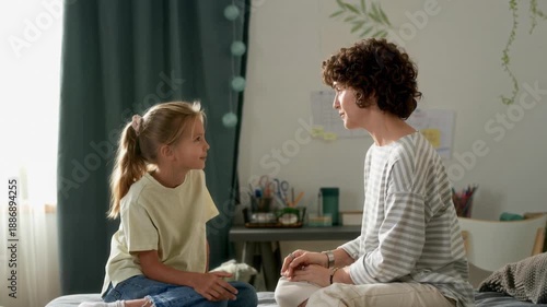 Side view of adult woman talking to cheerful young daughter discussing emotions together while sitting on bed at home, AI generated footage
