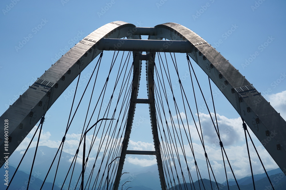 Obraz premium 青空を背にそびえ立つ巨大な鋼鉄製の橋のアーチ構造。連なる山々と白い雲を見下ろすようなダイナミックなアングルから捉えた近代的な土木建築の美しさ。