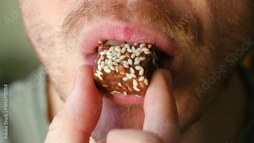 Bearded man eating healthy vegan energy bar covered in sesame seeds for quick and nutritious snack. Close up mouth chewing sweet candy, enjoying flavor. Man eating healthy sesame seed energy bar