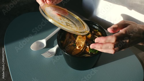 Woman hands opening plastic takeout container of hot asian noodle soup on modern table, with sunlight casting dramatic shadows, revealing ready to eat meal for convenient lunch. Asian noodle soup