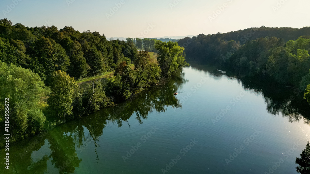 Obraz premium Aerial view of the Kolpa River near Metlika, Slovenia, showing a small river weir, calm green water, surrounding forest, and riverside recreation in a peaceful rural landscape during summer.