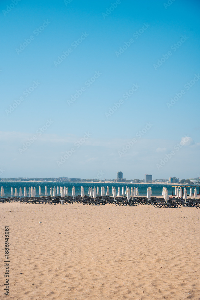 Fototapeta premium Visitors relax on sunbeds with umbrellas at a beach in Bulgaria. The coastline of sunny beach stretches along the water, while buildings rise in the distance. It is daytime and the sky is clear