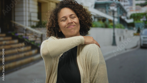 Photos Woman touching her shoulder with closed eyes on city street near stone building steps while wearing knit cardigan; relief