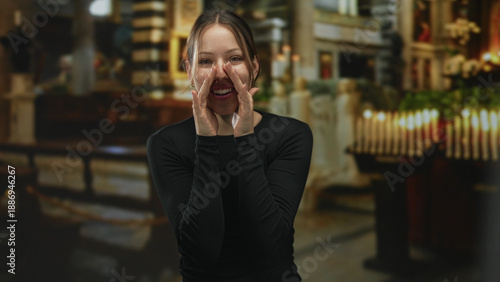 Slika na platnu Woman with hands cupped to mouth calling aloud in a catholic church building by lit votive candles and wooden pews, facing forward; joyful praise