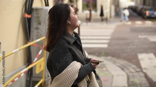 Side view of a woman standing on an urban street wearing wireless earbuds and talking. She is wearing a grey jacket and a patterned scarf draped over her arm.