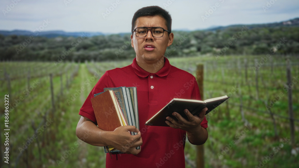 Fototapeta premium Young man wearing red polo and glasses holding several books and reading from an open notebook in a forest vineyard; study concentration.